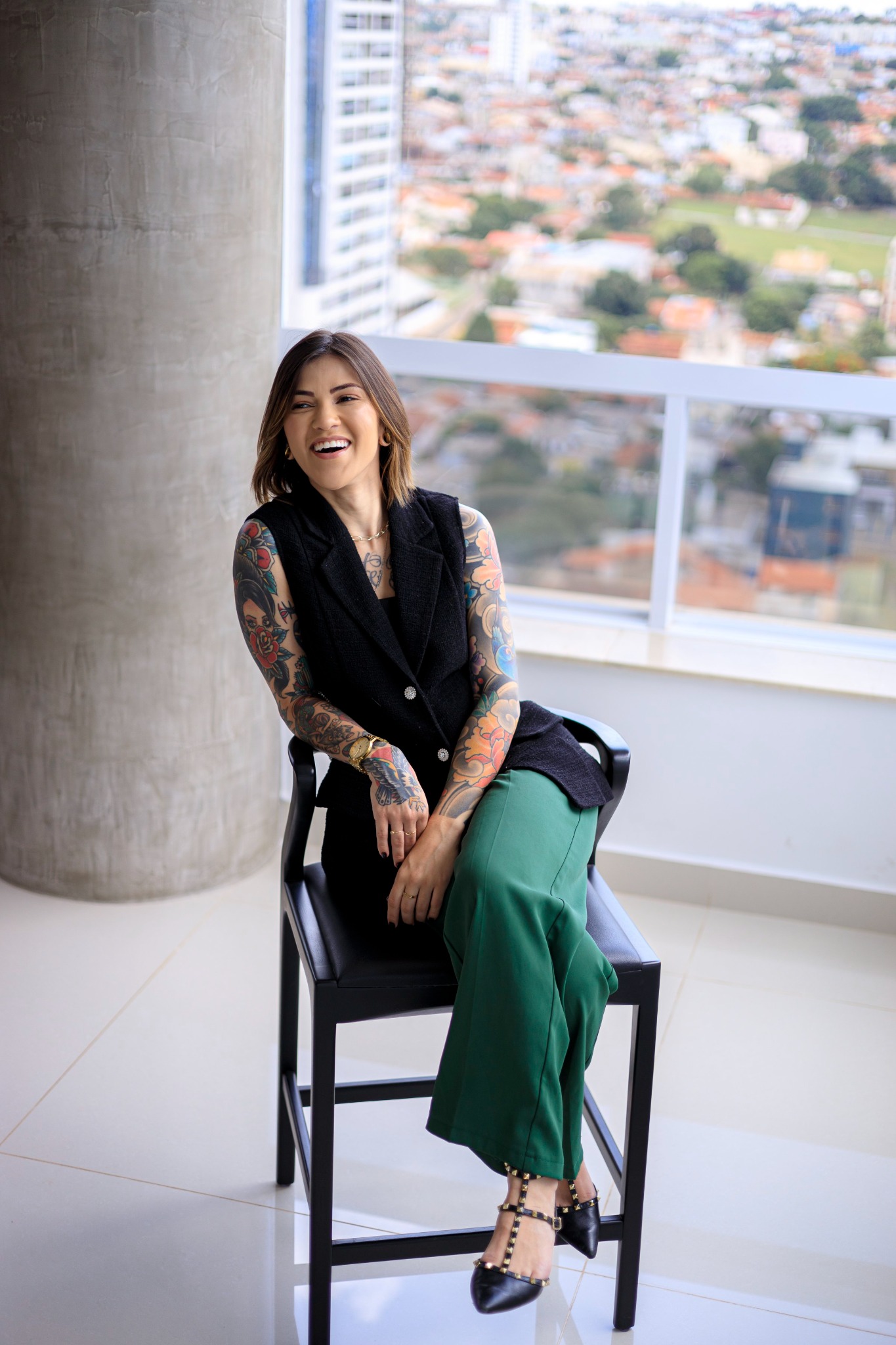 Dra. Thaís Ferrera, com roupa preta e verde, sorrindo junto à janela